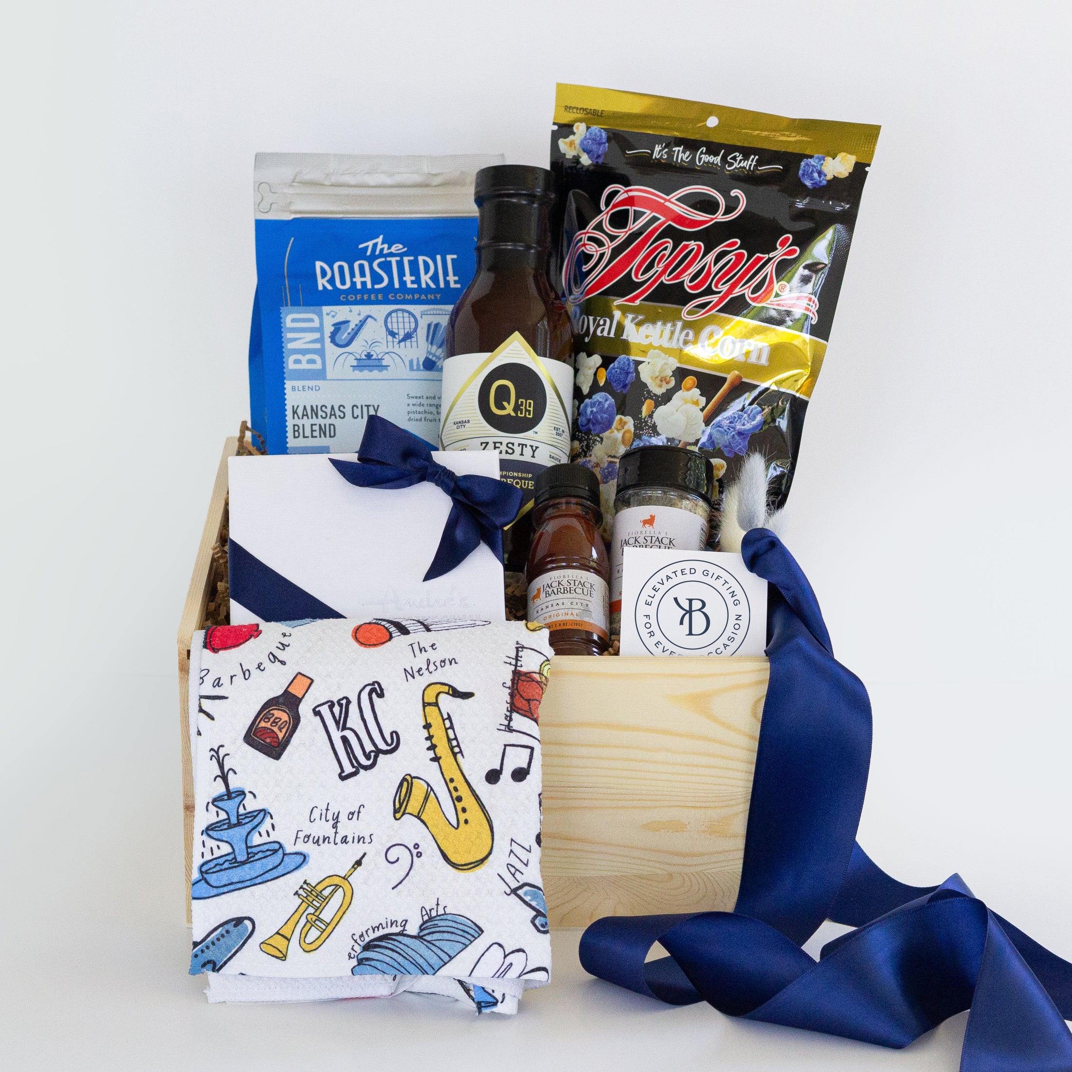 A curated gift basket in a light wood crate filled with assorted items, including a Kansas City themed kitchen towel, a blue bag of The Roasterie coffee, a bag of Topsy's Popcorn in Kansas City Royals blue kettle corn mix, a bottle of Q39 Barbecue Sauce, mini bottles of Jack Stack BBQ Sauce and All Purpose Seasoning, and a white assorted chocolates gift box with a blue ribbon, all arranged on a white background.