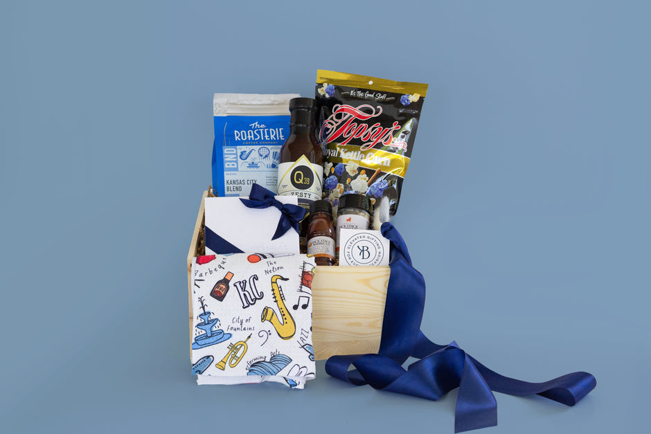 A curated gift basket in a light wood crate filled with assorted items, including a Kansas City themed kitchen towel, a blue bag of The Roasterie coffee, a bag of Topsy's Popcorn in Kansas City Royals blue kettle corn mix, a bottle of Q39 Barbecue Sauce, mini bottles of Jack Stack BBQ Sauce and All Purpose Seasoning, and a white assorted chocolates gift box with a blue ribbon, all arranged on a Bow on Top brand-accurate mist blue background.