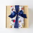 Load image into Gallery viewer, A gift box wrapped in a white Kansas City themed kitchen towel with a colorful illustrated pattern, tied with a large navy blue satin ribbon, presented in a light wood box on a white background.
