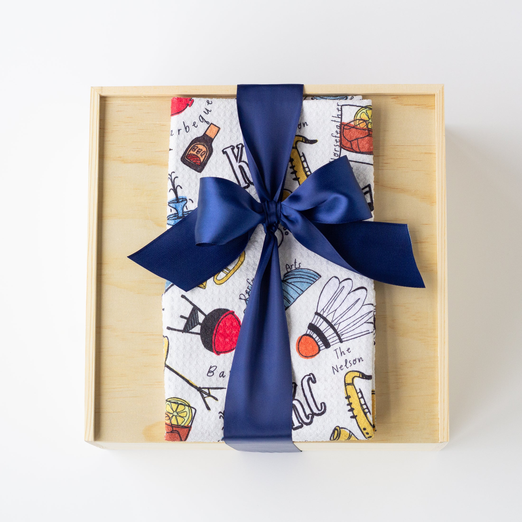 A gift box wrapped in a white Kansas City themed kitchen towel with a colorful illustrated pattern, tied with a large navy blue satin ribbon, presented in a light wood box on a white background.