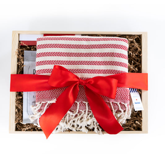 A folded red and white patterned turkish linen presented in a light wood gift box with shredded paper filler and tied with a wide red ribbon. A small tag is visible on the right side.
