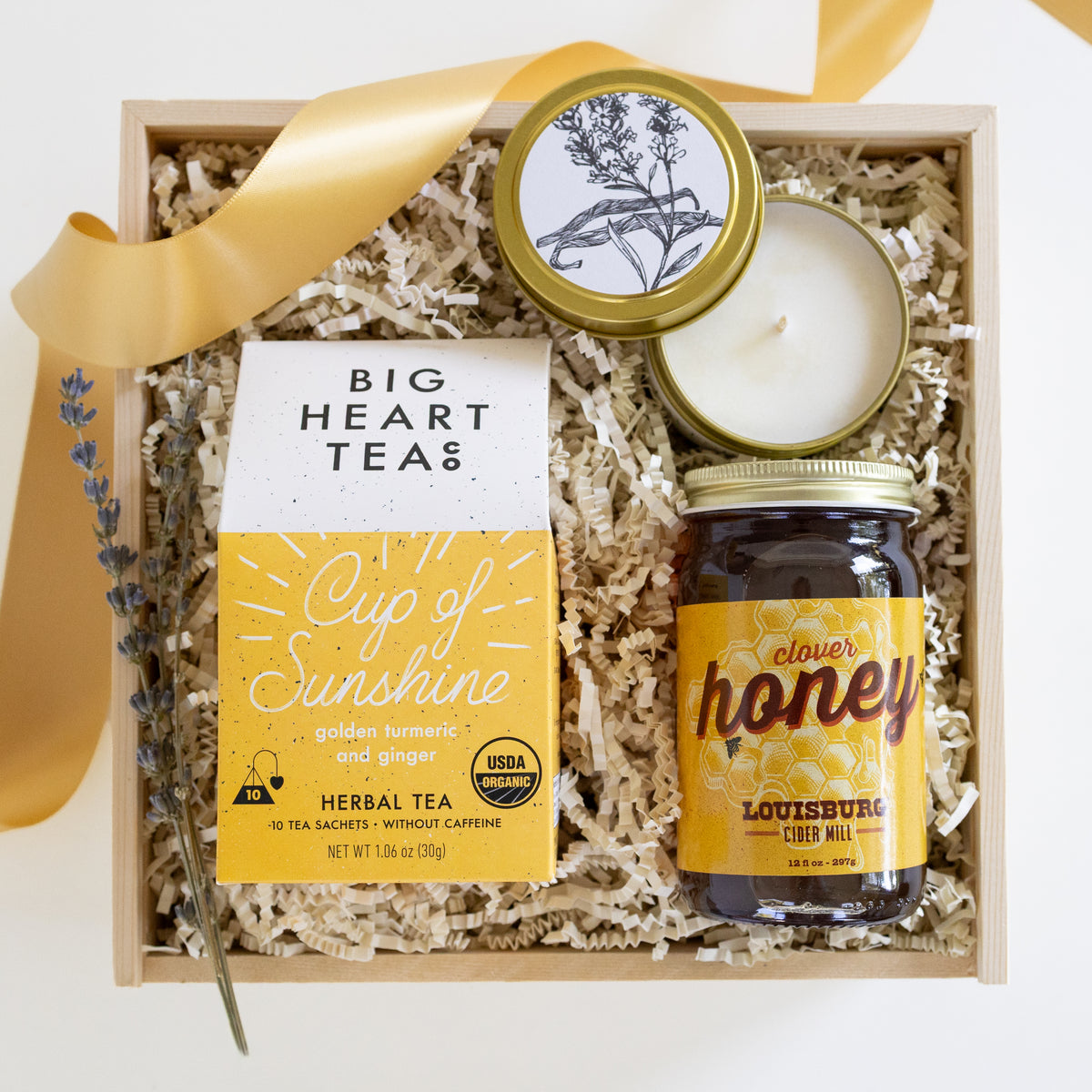 A gift box set containing Big Heart Tea's 'Cup of Sunshine' herbal tea, Louisburg's Clover Honey, a candle, and lavender sprigs in a wooden tray with shredded paper fill and a gold ribbon.