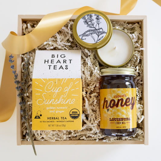 A gift box set containing Big Heart Tea's 'Cup of Sunshine' herbal tea, Louisburg's Clover Honey, a candle, and lavender sprigs in a wooden tray with shredded paper fill and a gold ribbon.