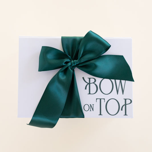 A white rectangular box tied with a large satin bow in deep teal green, featuring the printed phrase 'BOW ON TOP' in dark green lettering on the front, set against a light cream background.
