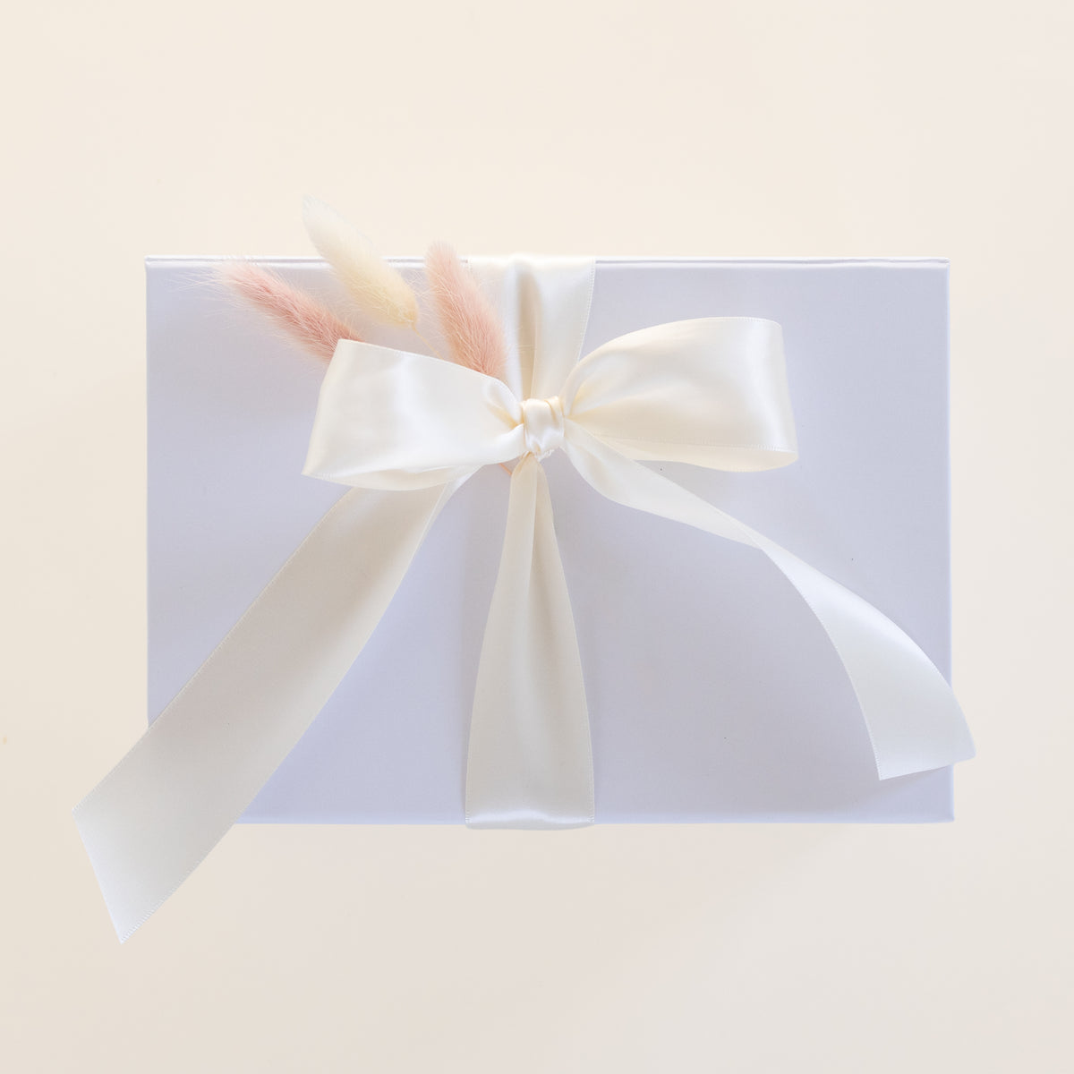 A square white gift box wrapped with a large cream satin ribbon bow and tied with dried pastel grass stems, photographed on a warm off-white background.