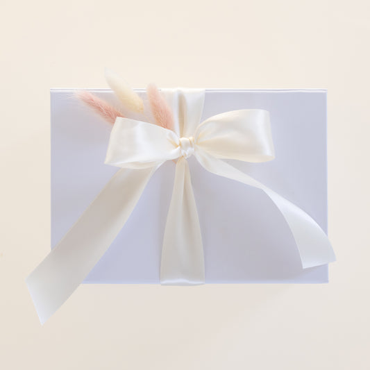 A square white gift box wrapped with a large cream satin ribbon bow and tied with dried pastel grass stems, photographed on a warm off-white background.