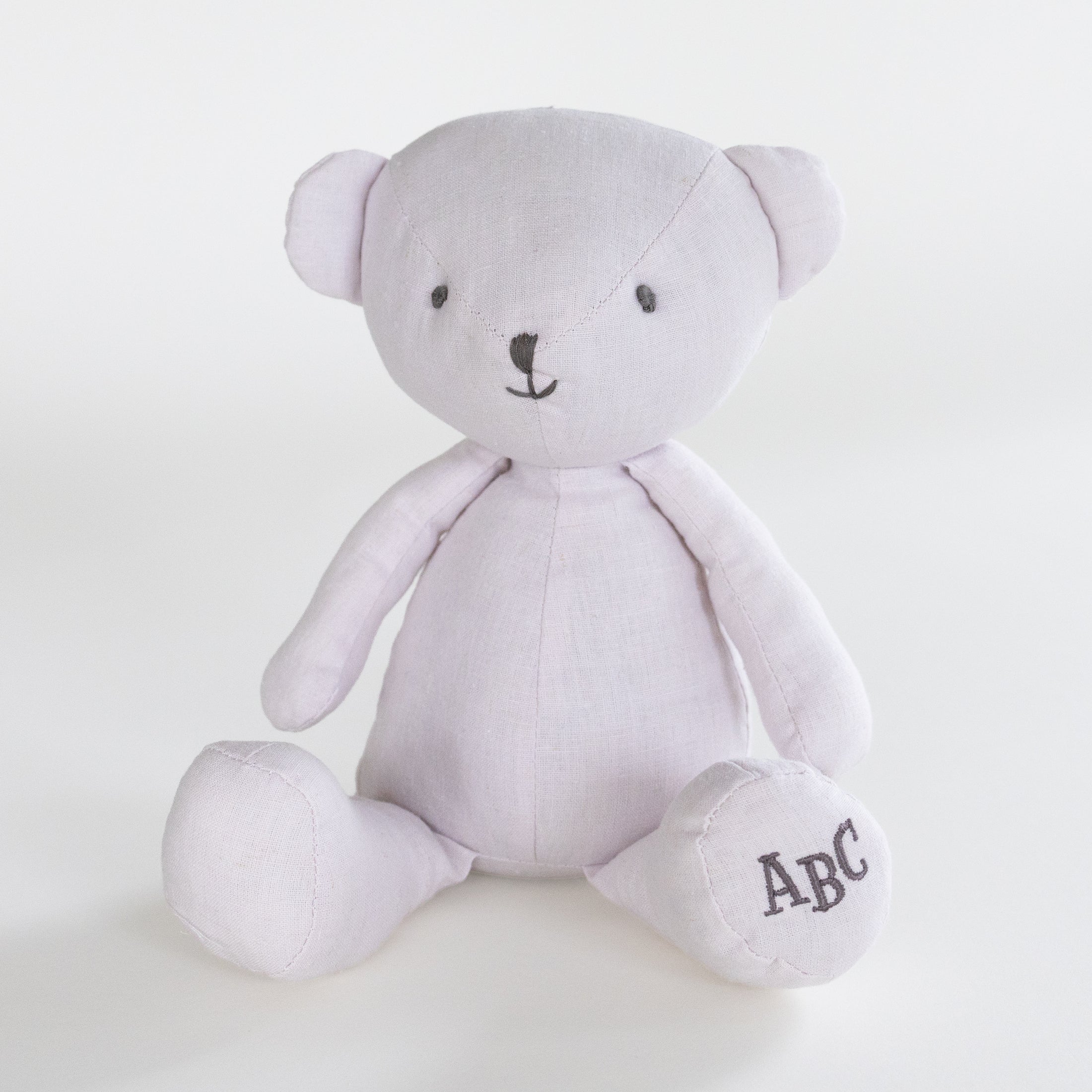 A light lavender plush teddy bear with black embroidered facial features and the letters 'ABC' stitched on its right foot, set against a plain white background.