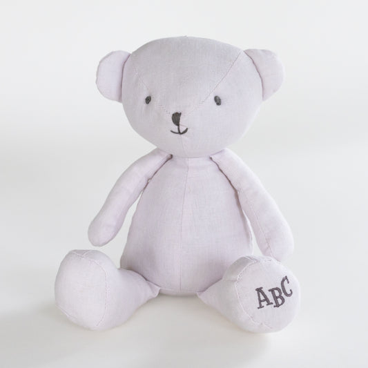 A light lavender plush teddy bear with black embroidered facial features and the letters 'ABC' stitched on its right foot, set against a plain white background.