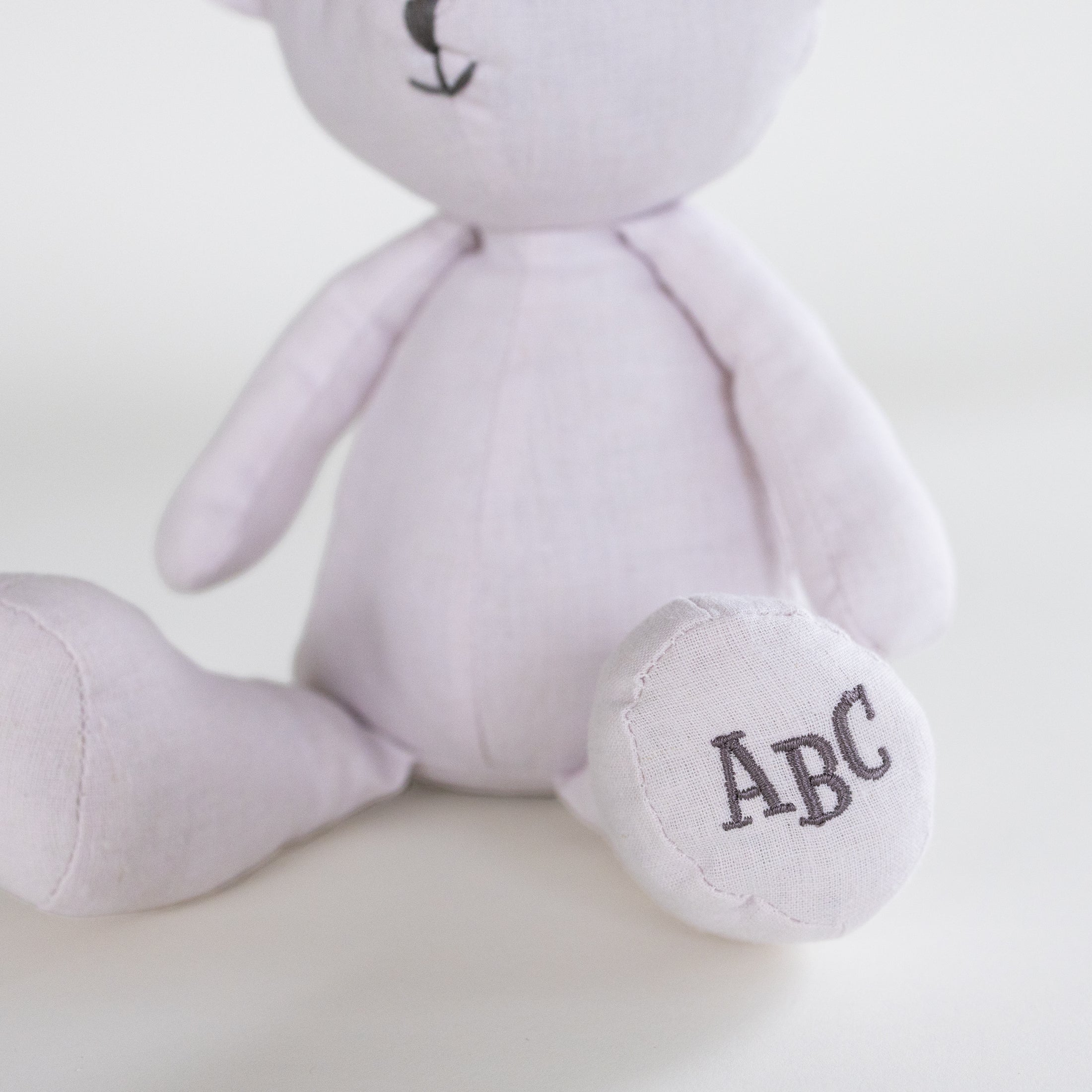 A close-up of a light lavender plush teddy bear sitting down, with its foot customized with the embroidered initials 'ABC' in dark gray.
