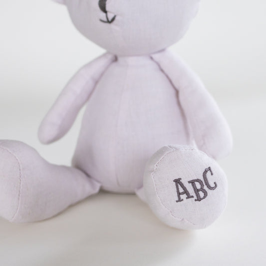 A close-up of a light lavender plush teddy bear sitting down, with its foot customized with the embroidered initials 'ABC' in dark gray.