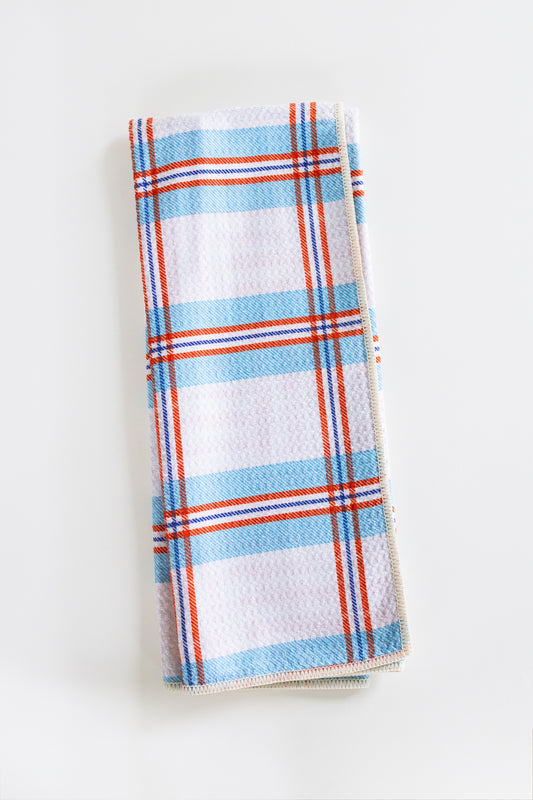 A folded kitchen towel with a white waffle-weave texture, featuring a plaid pattern of light blue, orange, and thin red stripes, set against a plain white background.