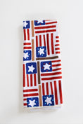Load image into Gallery viewer, A folded white kitchen towel featuring a repeating patriotic American flag pattern with red stripes, blue fields, and white stars, displayed on a clean white background.

