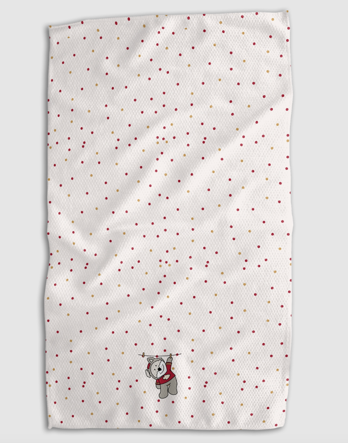 A white textured tea towel with a scattered pattern of small red and gold dots, featuring a small gray wolf plushie wearing a red sweater near the bottom center, shown against a neutral gray background.