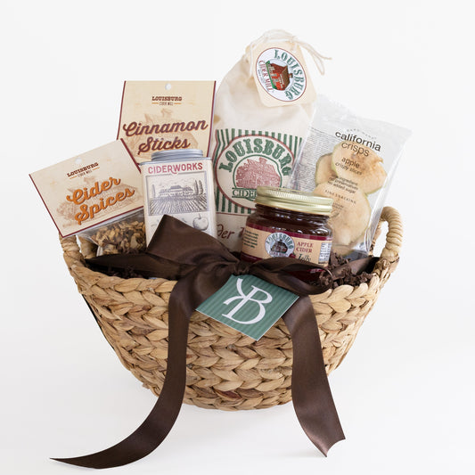 A woven straw gift basket filled with assorted gourmet snacks and treats, including a jar of Louisburg Apple Cider Jelly, cinnamon sticks, cider spices, and California Apple Crisps, tied with a dark brown ribbon and a green monogram tag, on a white background.