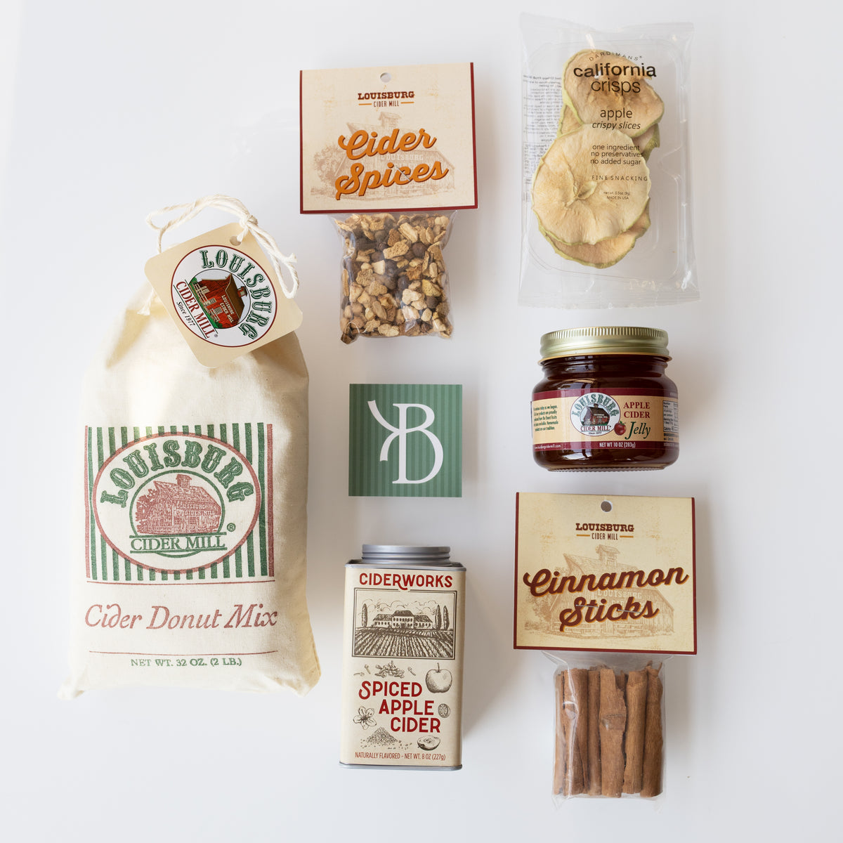 A flat-lay arrangement of Louisburg Ciderworks gift items on a white background, including a large bag of cider donut mix, a jar of apple cider, cinnamon sticks, apple slices, spices, and a tin of spiced apple cider.