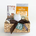 Load image into Gallery viewer, A woven natural fiber gift basket tied with a dark brown ribbon and a green monogram tag, filled with assorted gift items including a chocolate bar with the Eiffel Tower, a jar candle, and a bag of coffee from The Roasterie photographed on a clean white background.
