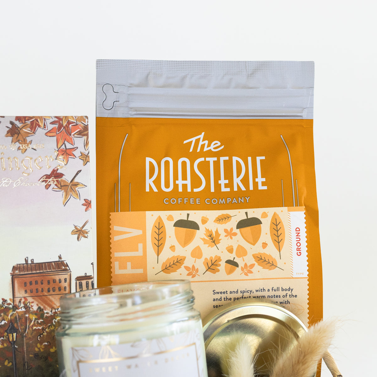 A coffee bag from The Roasterie Coffee Company stands upright on a white background, styled with a glass jar candle, a metal tin lid, and a chocolate bar label featuring autumn leaves and a building illustration.