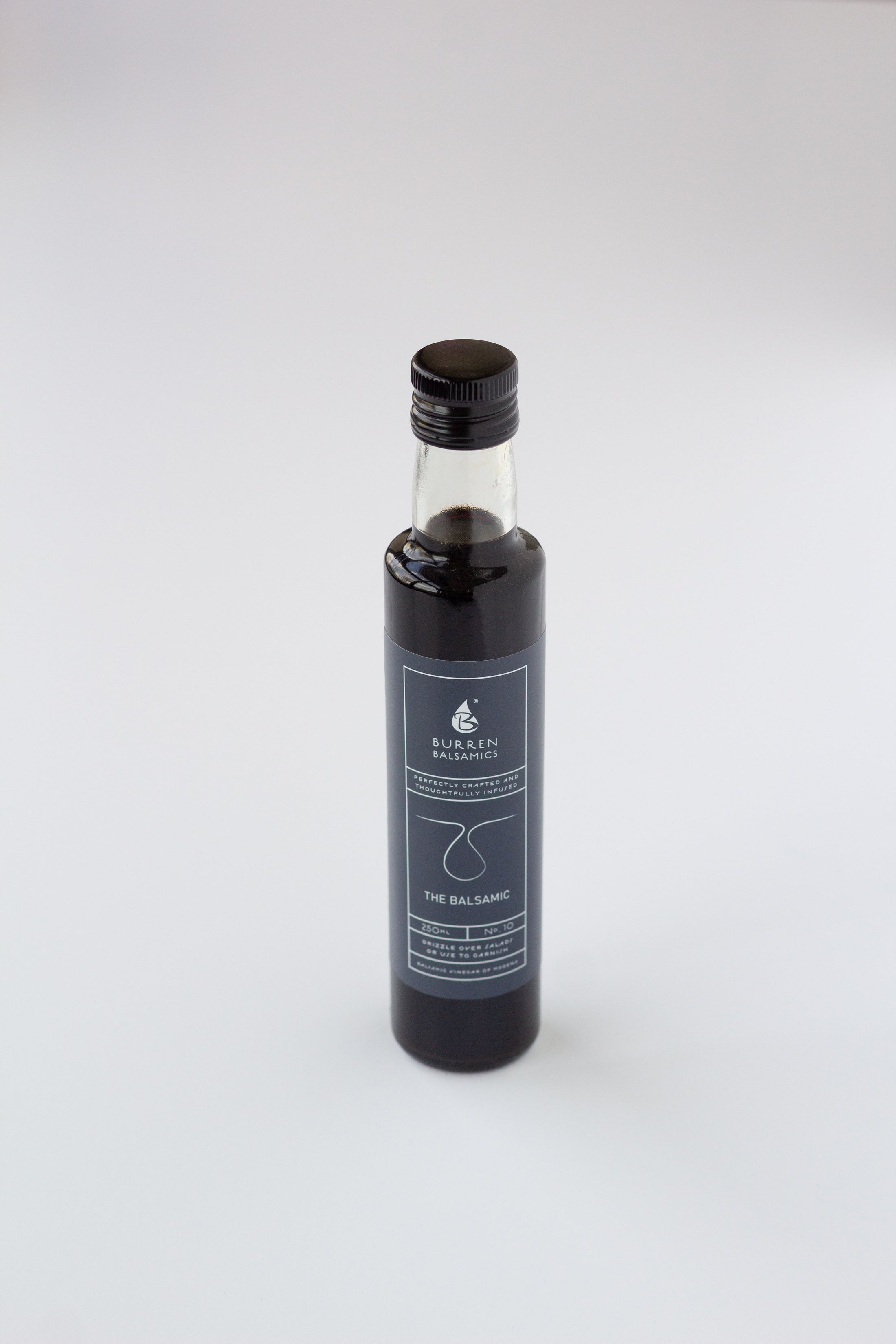 A glass bottle of balsamic vinegar with a black cap and a dark blue label, standing upright on a light gray background.