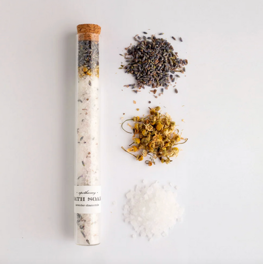 A bath soak product shown in a clear glass tube with a cork stopper, alongside loose dried lavender, chamomile flowers, and coarse salt crystals on a white background. The tube label reads 'Apothecary Bath Soak Lavender Chamomile'