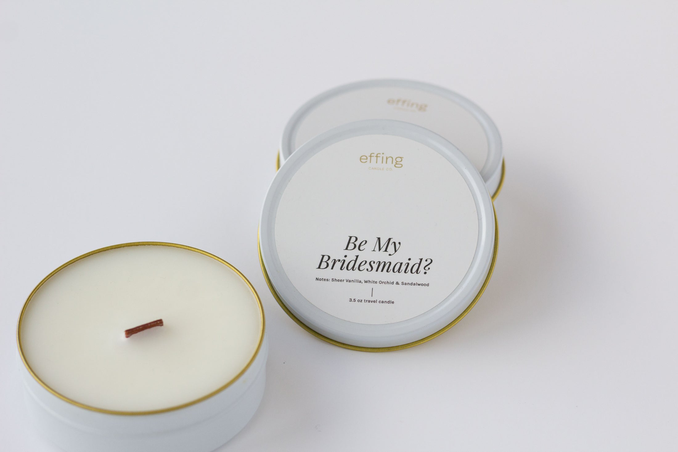 Two round candles in white tins with gold rims on a clean white background. One tin is open, showing a white wax surface and a small brown wood wick.