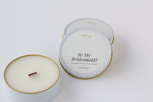 Two round candles in white tins with gold rims on a clean white background. One tin is open, showing a white wax surface and a small brown wood wick.