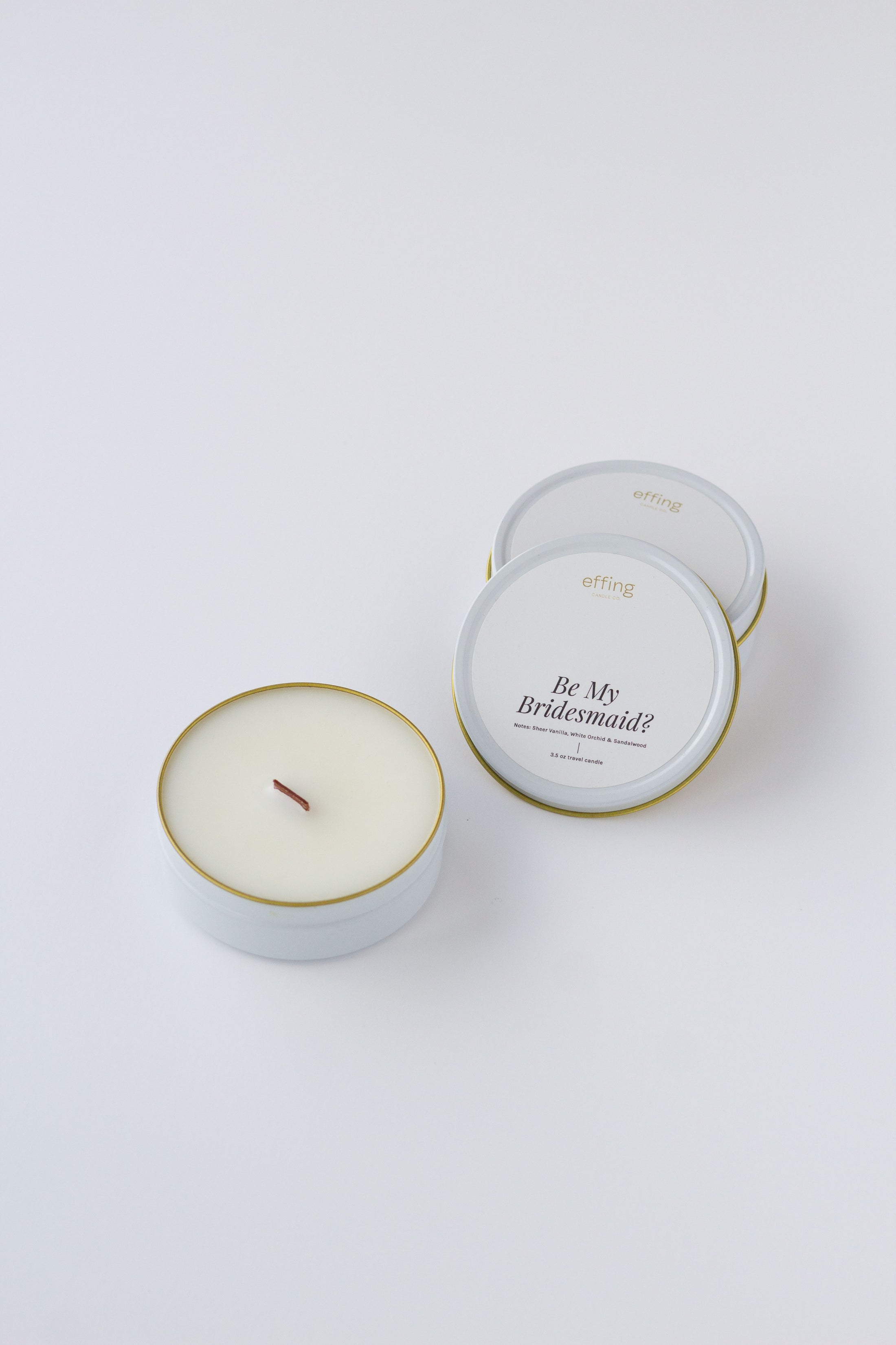 A white wax candle in a round white tin with a thin gold rim, shown next to one closed matching tin on a clean white background. One open tin label reads 'Be My Bridesmaid' and the brand 'effing candle co' is visible at the top.