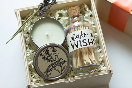 A birthday gift set in a light wood box filled with white paper shred, featuring a white candle in a silver tin with a botanical bird illustration., and a small glass bottle of matches labeled 'make a WISH!' with a cork stopper.