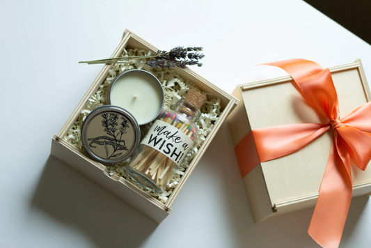 A birthday gift set in a light wood box filled with white paper shred, featuring a white candle in a silver tin with a botanical bird illustration., and a small glass bottle of matches labeled 'make a WISH!' with a cork stopper.