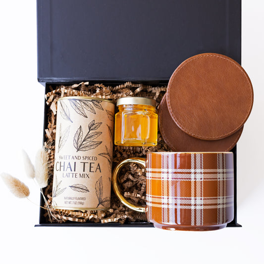 A curated gift set in an open black box filled with crinkle paper. The set includes a canister of Sweet and Spiced Chai Tea Latte Mix, a small glass jar of golden honey, a brown leather coaster set, and a brown plaid ceramic mug.