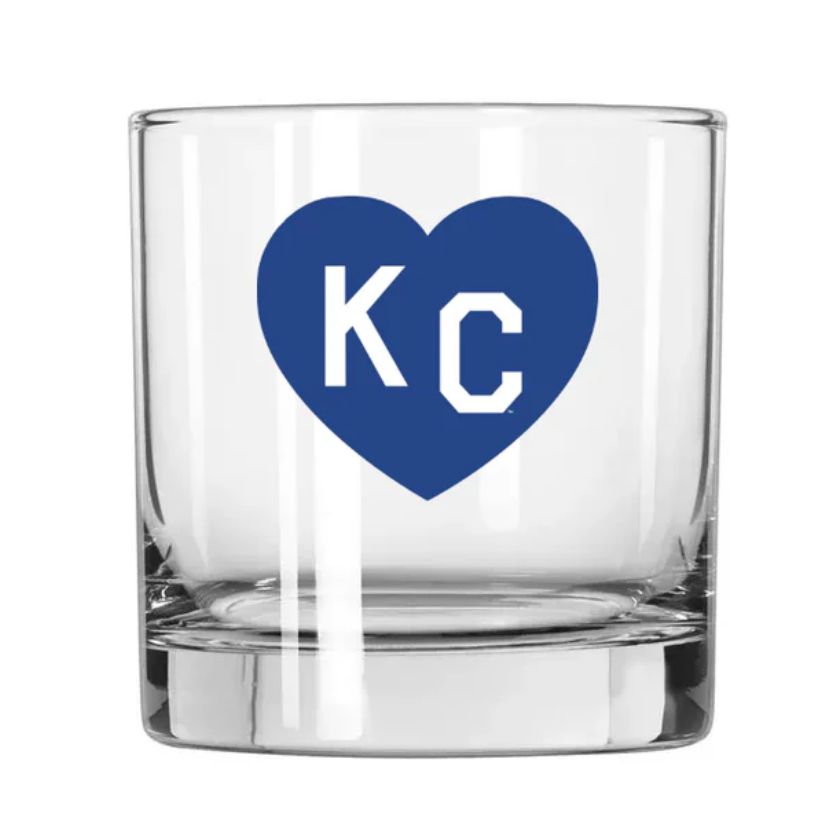A clear glass rocks tumbler featuring a blue heart graphic on the front with the white letters 'KC' inside, set against a plain white background.