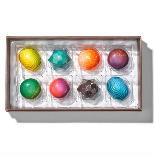 A boxed assortment of colorful chocolates in a clear tray with a metallic brown rim, photographed from above on a white background with a soft shadow.