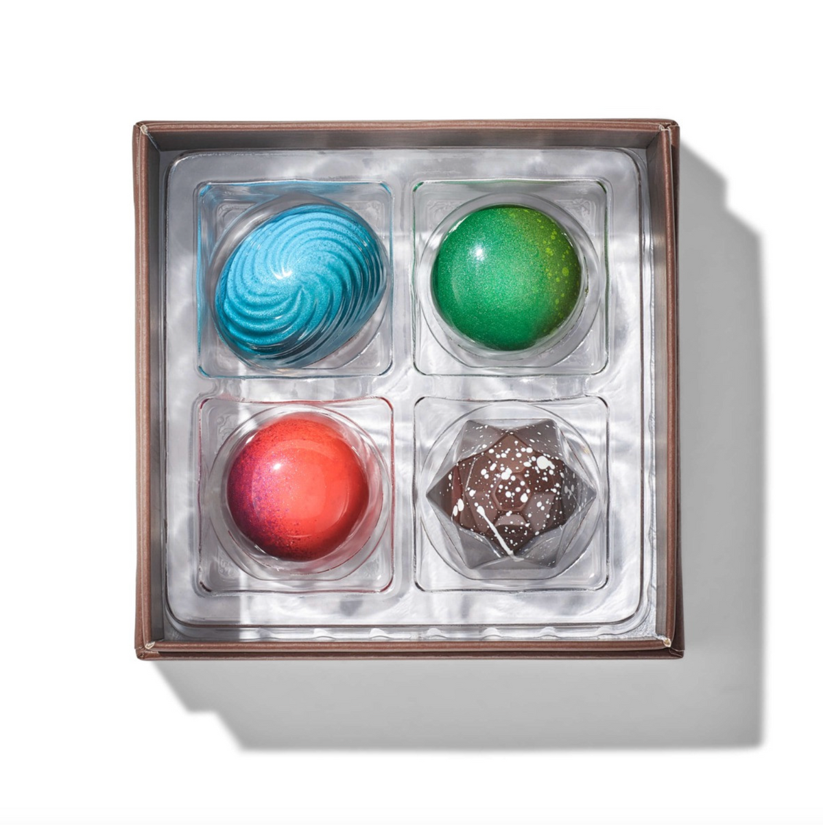 A boxed assortment of four gourmet chocolates in a square brown box with clear plastic compartments, photographed from above on a white background with a soft shadow.