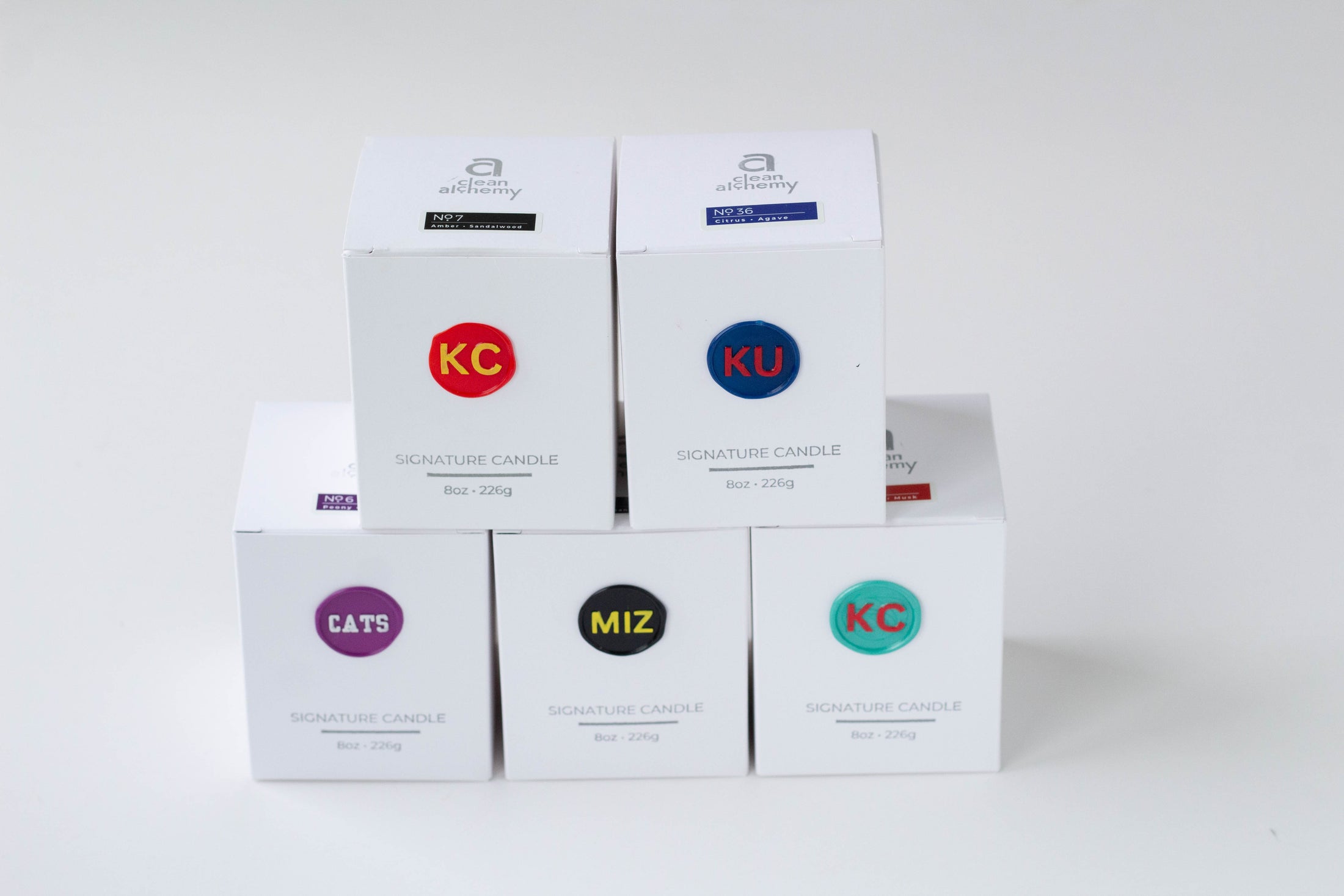 Five white product boxes arranged in a small stack on a light gray background. The boxes feature colorful circular logo labels with text including KC, KU, CATS, and MIZ, along with the words SIGNATURE CANDLE.
