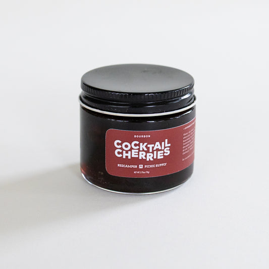 A small glass jar of Bourbon Cocktail Cherries with a black lid and a dark red label, set against a plain white background.