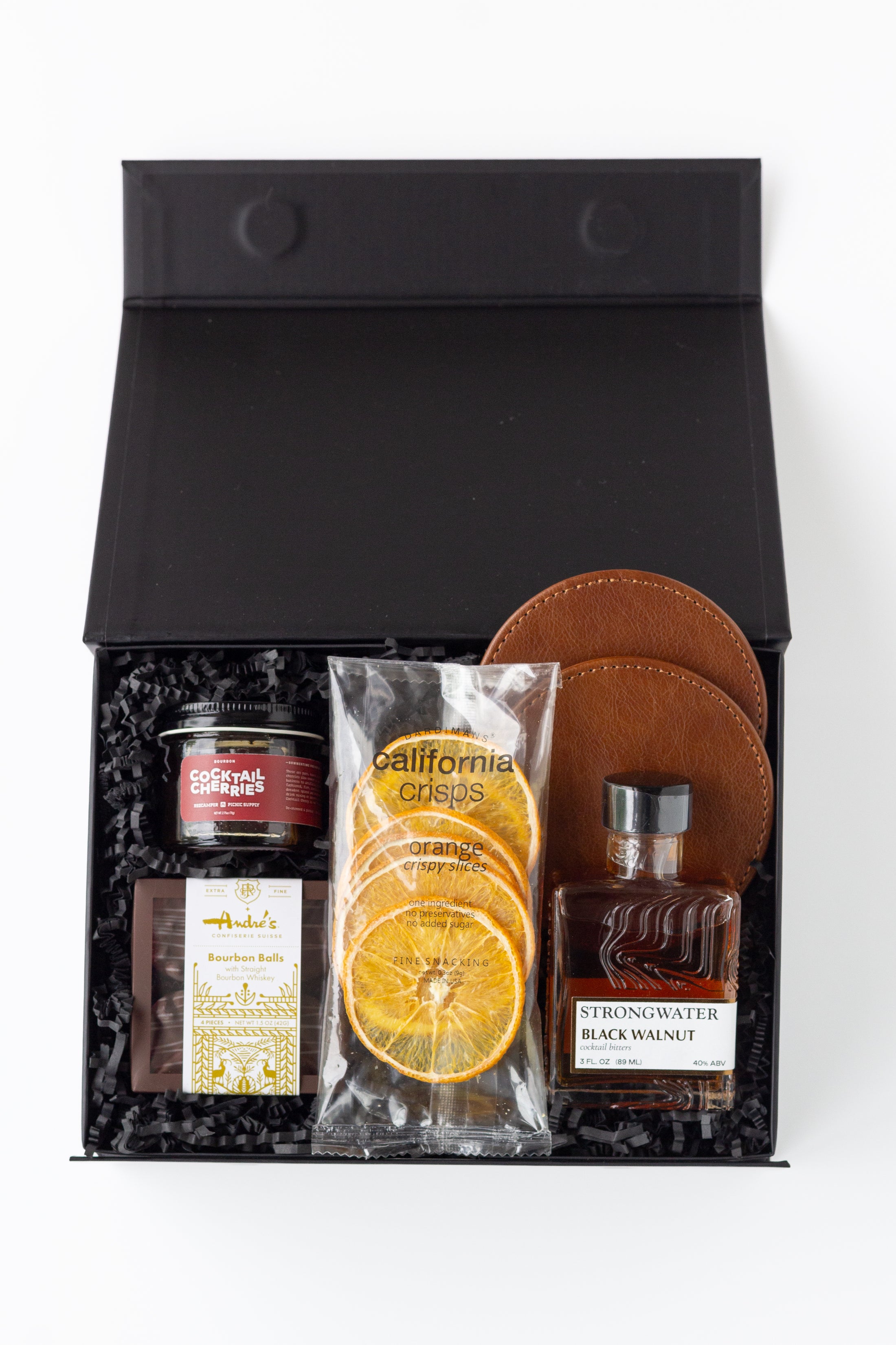 A curated gift box set in a black presentation box with shredded black filler, featuring a jar of cocktail cherries, a bag of California Crisps dried orange slices, a bottle of Strongwater Black Walnut cocktail bitters, a package of Andre's Bourbon Balls, and two brown leather coasters.
