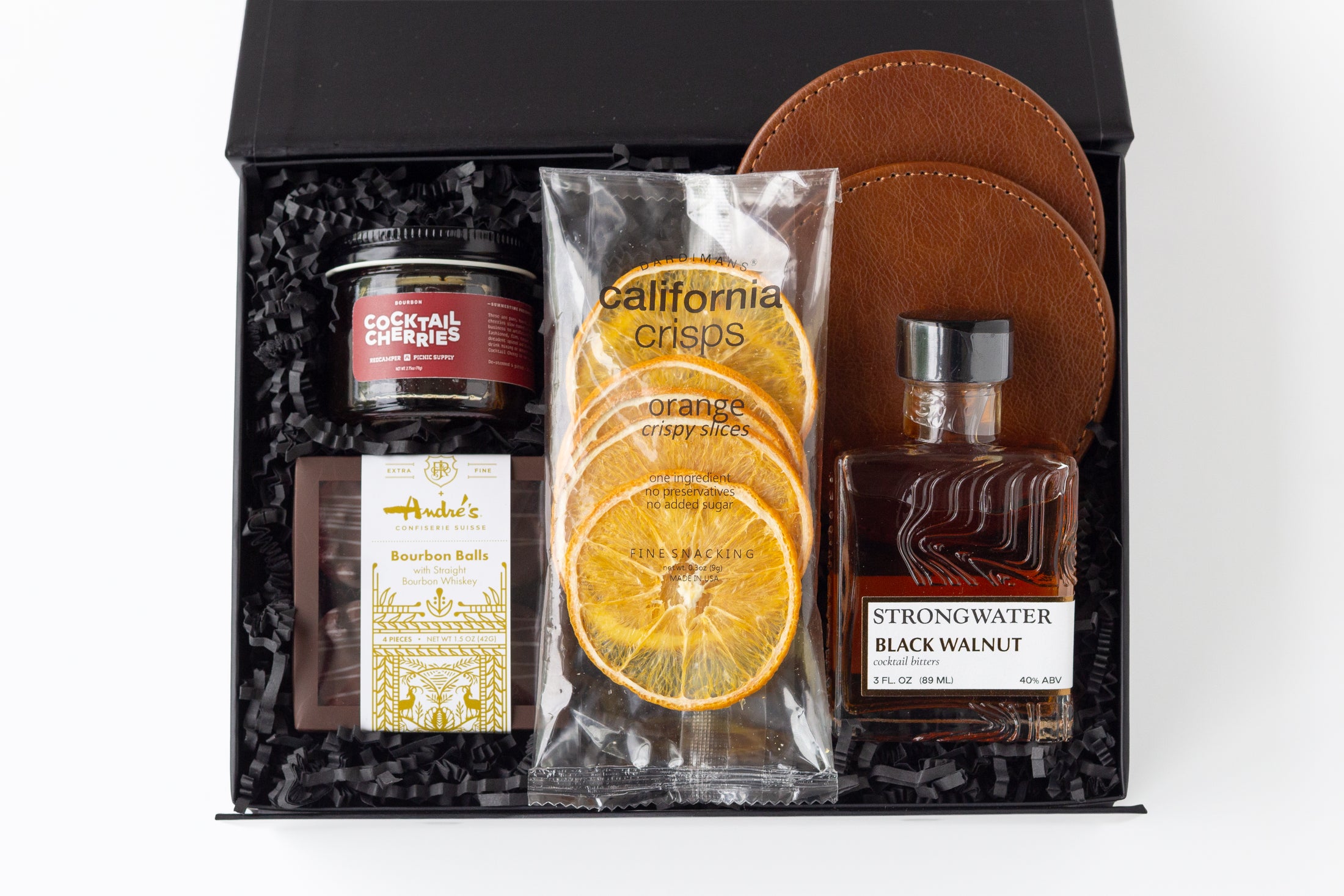 A curated gift box set in a black presentation box with shredded black filler, featuring a jar of cocktail cherries, a bag of California Crisps dried orange slices, a bottle of Strongwater Black Walnut cocktail bitters, a package of Andre's Bourbon Balls, and two brown leather coasters.