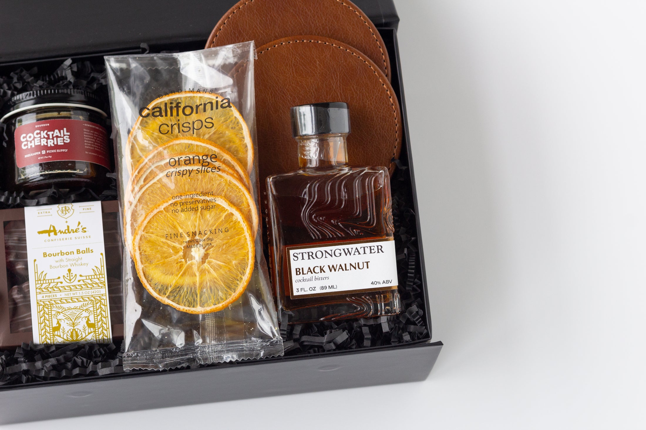 A curated gift box set in a black presentation box with shredded black filler, featuring a jar of cocktail cherries, a bag of California Crisps dried orange slices, a bottle of Strongwater Black Walnut cocktail bitters, a package of Andre's Bourbon Balls, and two brown leather coasters.