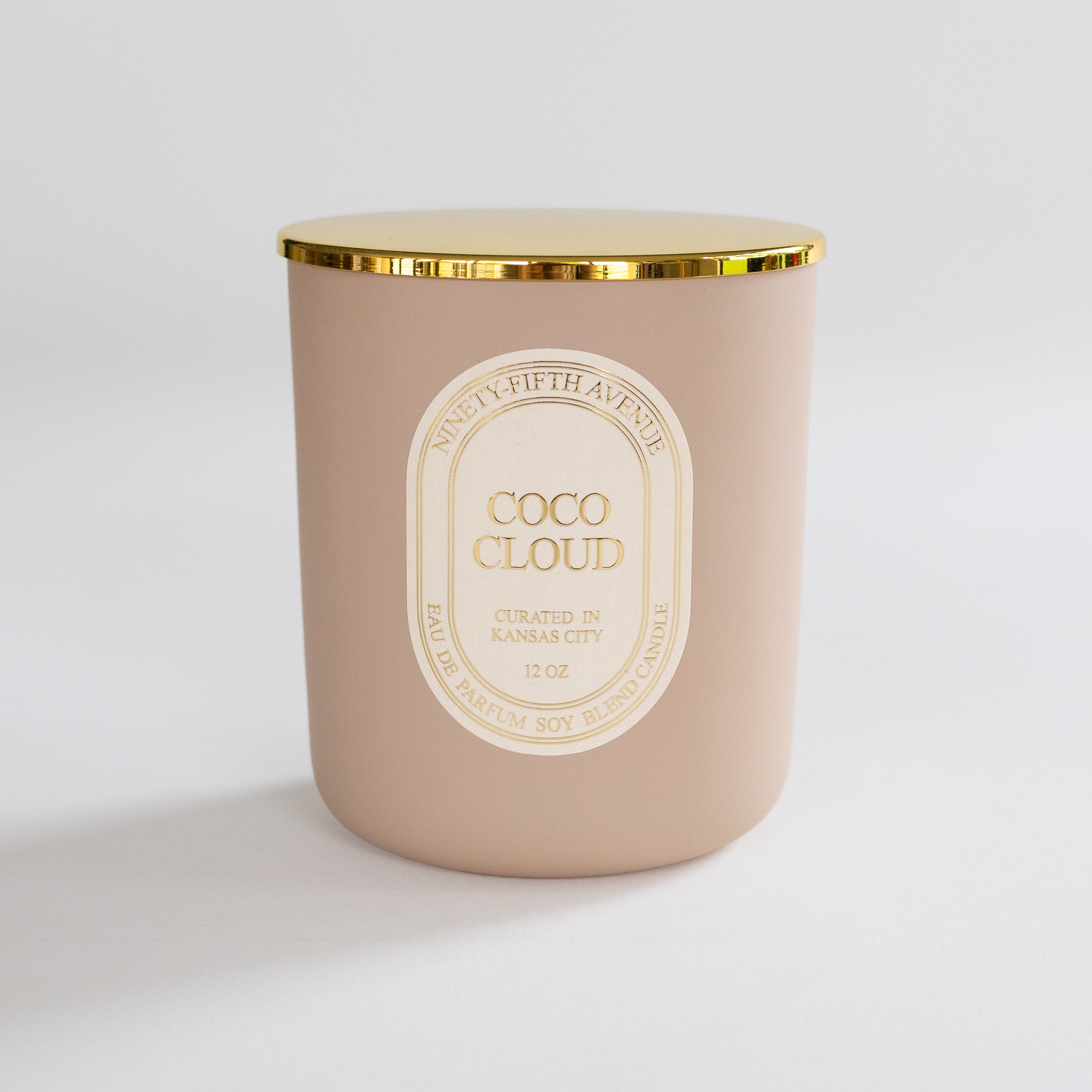 A matte blush pink candle jar with a gold metallic rim and a cream-colored oval label reading 'COCO CLOUD' and 'Ninety-Fifth Avenue.' The candle is photographed on a clean, light gray studio background.