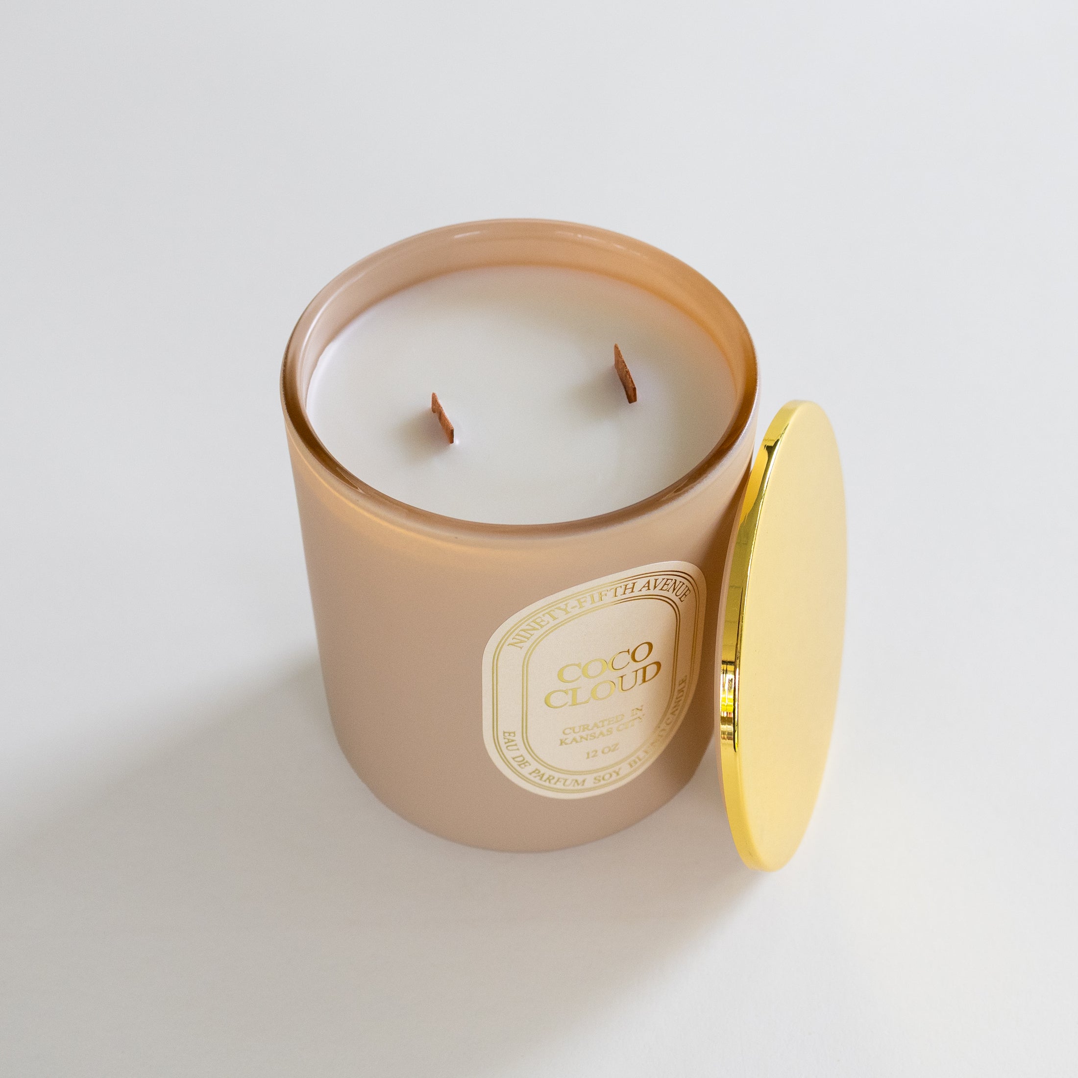 A candle in a frosted beige glass jar with a gold label reading 'COCO CLOUD', shown with its gold lid leaning against it on a clean white background.
