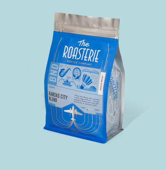 a blue Roasterie Kansas City Blend Whole Bean Coffee bag on a light blue background. The bag features iconic Kansas City symbols like the shuttlecock, various Kansas City buildings, a saxophone, a fountain, and the Roasterie plane logo. 