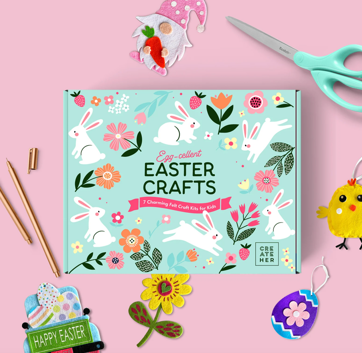 A flat-lay of an Easter craft kit box on a pink background, surrounded by themed props including scissors, wooden sticks, a gnome, a chick cutout, and painted Easter eggs. The box reads 'Easter Crafts' and shows white rabbits, flowers, and leaves on a light blue background.