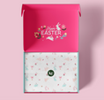 Load image into Gallery viewer, An open pink gift box with a floral 'Happy Easter' design on the inside lid, revealing a white tissue paper insert with a delicate bunny and flower pattern and a small green 'kc' logo. The box is set against a solid light pink background.

