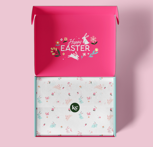 An open pink gift box with a floral 'Happy Easter' design on the inside lid, revealing a white tissue paper insert with a delicate bunny and flower pattern and a small green 'kc' logo. The box is set against a solid light pink background.