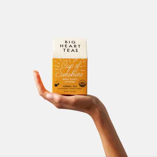 A hand holds a box of Cup of Sunshine turmeric herbal tea from Big Heart Tea company. Each box holds 10 tea bags.