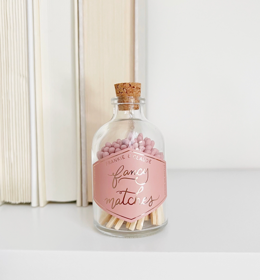 a glass jar of pink-tipped matches on a bookshelf ledge. The label on the glass jar reads "Fancy Matches"