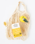 Load image into Gallery viewer, A cream-colored mesh net farmer's market bag filled with Big Heart Tea's 'Cup of Sunshine' herbal tea, styled with a yellow lemon-shaped candle and a small wrapped honeycomb lollipop on a clean white background.
