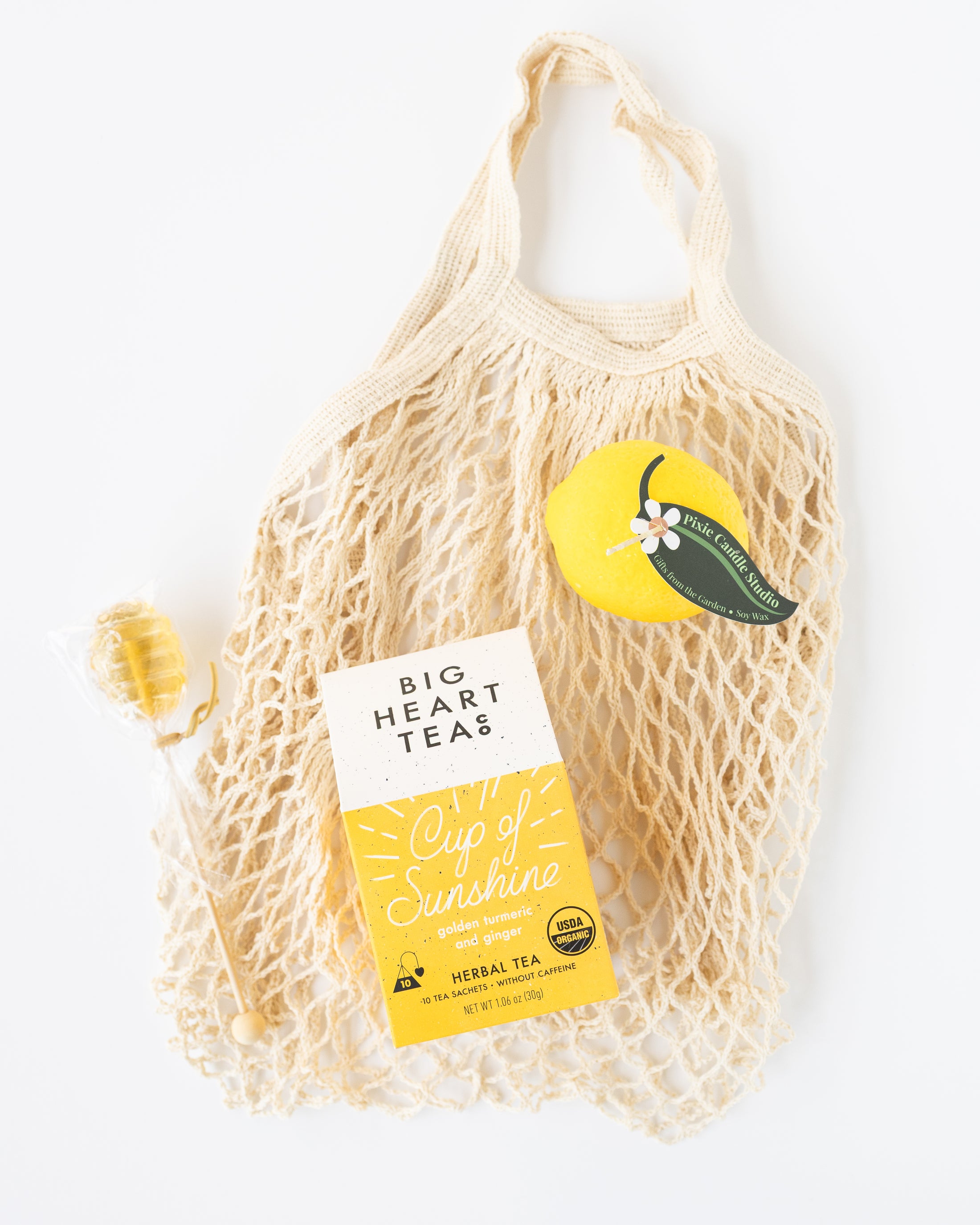 A cream-colored mesh net farmer's market bag filled with Big Heart Tea's 'Cup of Sunshine' herbal tea, styled with a yellow lemon-shaped candle and a small wrapped honeycomb lollipop on a clean white background.