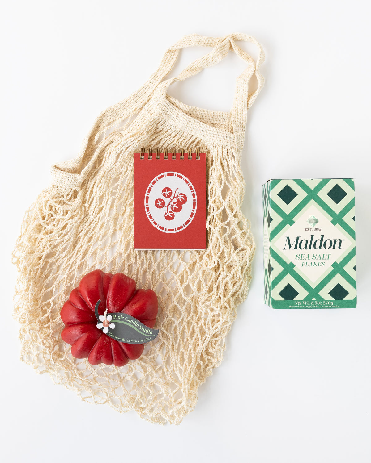 A Farmer's Market-themed gift with a mesh net bag, a tomato-shaped red candle, a red mini notebook with a tomato graphic on top, and a box of Maldon salt just out of frame.