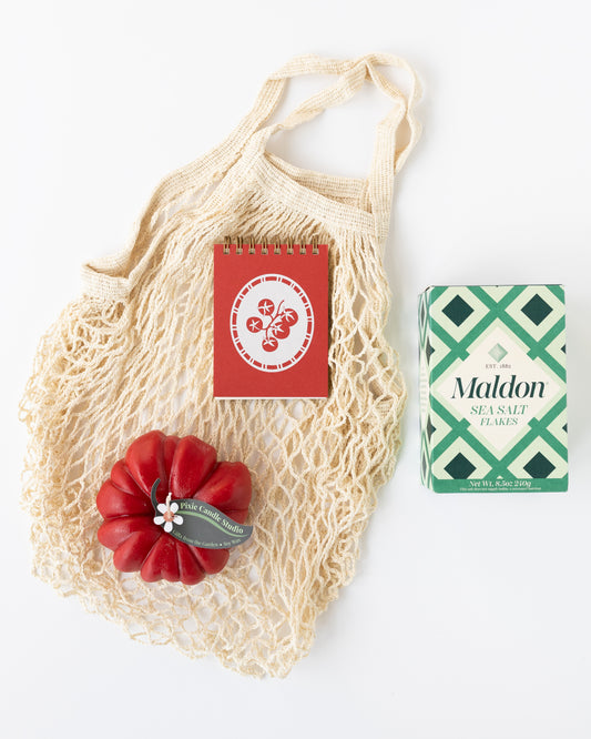 A Farmer's Market-themed gift with a mesh net bag, a tomato-shaped red candle, a red mini notebook with a tomato graphic on top, and a box of Maldon salt just out of frame.