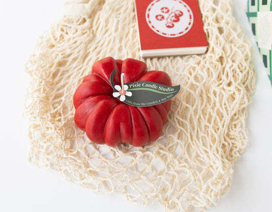 A Farmer's Market-themed gift with a mesh net bag, a tomato-shaped red candle, a red mini notebook with a tomato graphic on top, and a box of Maldon salt just out of frame.